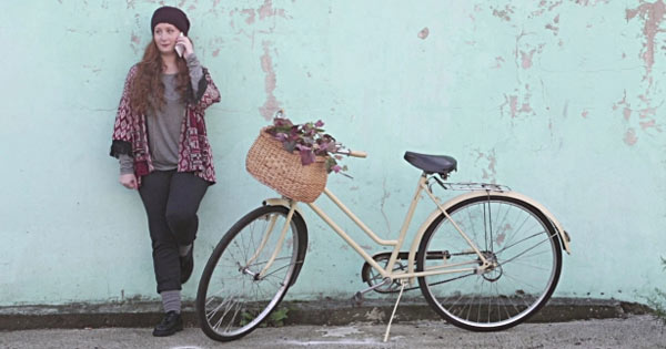 Bellende jonge vrouw leunt tegen een groene muur. Voor haar een witte damesfiets met fietsmand.
